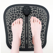 Top-down view of EMS foot massager pad with real feet positioned on surface, showing geometric grid pattern and black wireless control unit