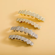 Four iced-out tooth grillz pieces laid flat on warm beige background showing two gold and two silver rhinestone-paved sets