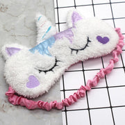 Two white fluffy cat unicorn sleep masks with purple heart embroidery, blue and purple satin accents, and pink ruched elastic bands displayed on marble and grid surface