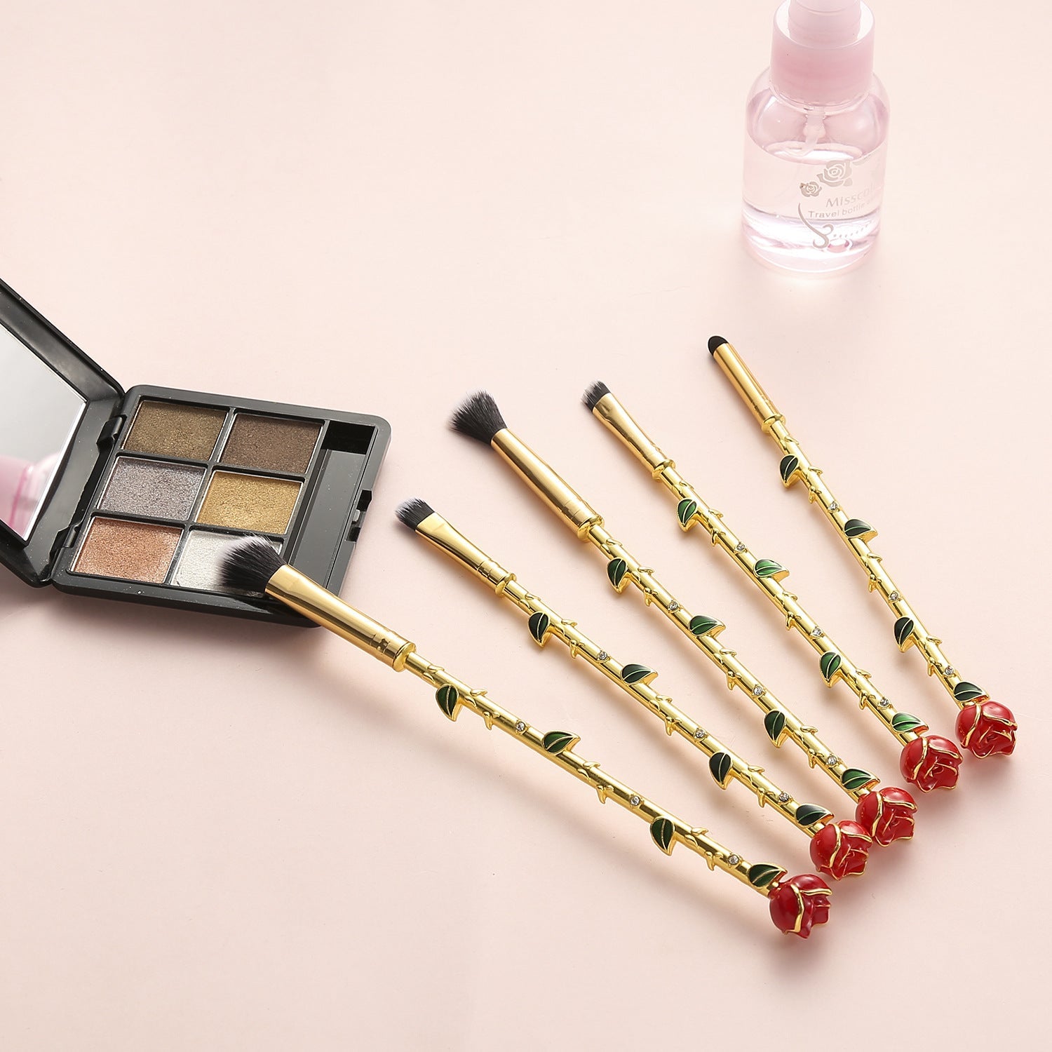 Five rose-stem makeup brushes fanned out beside an open shimmer eyeshadow palette and small pink bottle on a blush pink background