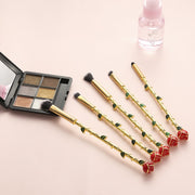 Five rose-stem makeup brushes fanned out beside an open shimmer eyeshadow palette and small pink bottle on a blush pink background