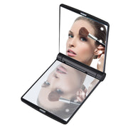 Black folding LED compact mirror open at 90 degrees reflecting a woman applying makeup with a brush, LED lights visible glowing on upper panel