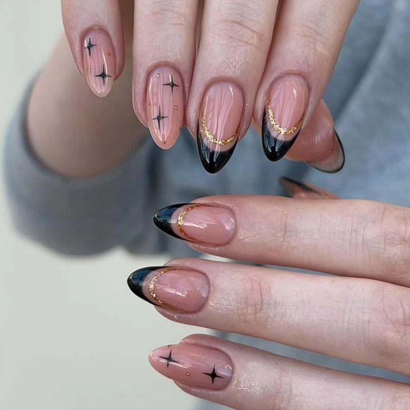 Close-up of two hands wearing almond-shaped press-on nails with black French tips, gold glitter crescent accents, and black sparkle star motifs on a nude blush base