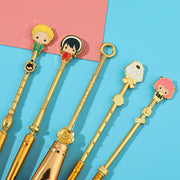 Five gold anime makeup brushes displayed flat on a turquoise and pink surface showing enamel character charms on each handle top