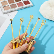 Hand holding a fan of 7 gold One Piece anime makeup brushes with character charms, displayed against a blue background with an eyeshadow palette in the background