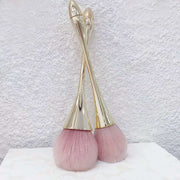 Two gold kabuki powder brushes with blush pink synthetic bristles crossed elegantly against a white textured background