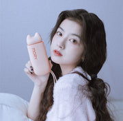 Young Asian woman in fluffy pink top holding peach-colored MORY cat mini fascia gun up near her face against lavender-grey background