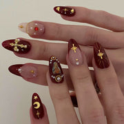 Two hands wearing elaborate dark burgundy press-on nails with gold cross charms, pearl accents, celestial motifs, and a Virgin Mary icon decal against a neutral background
