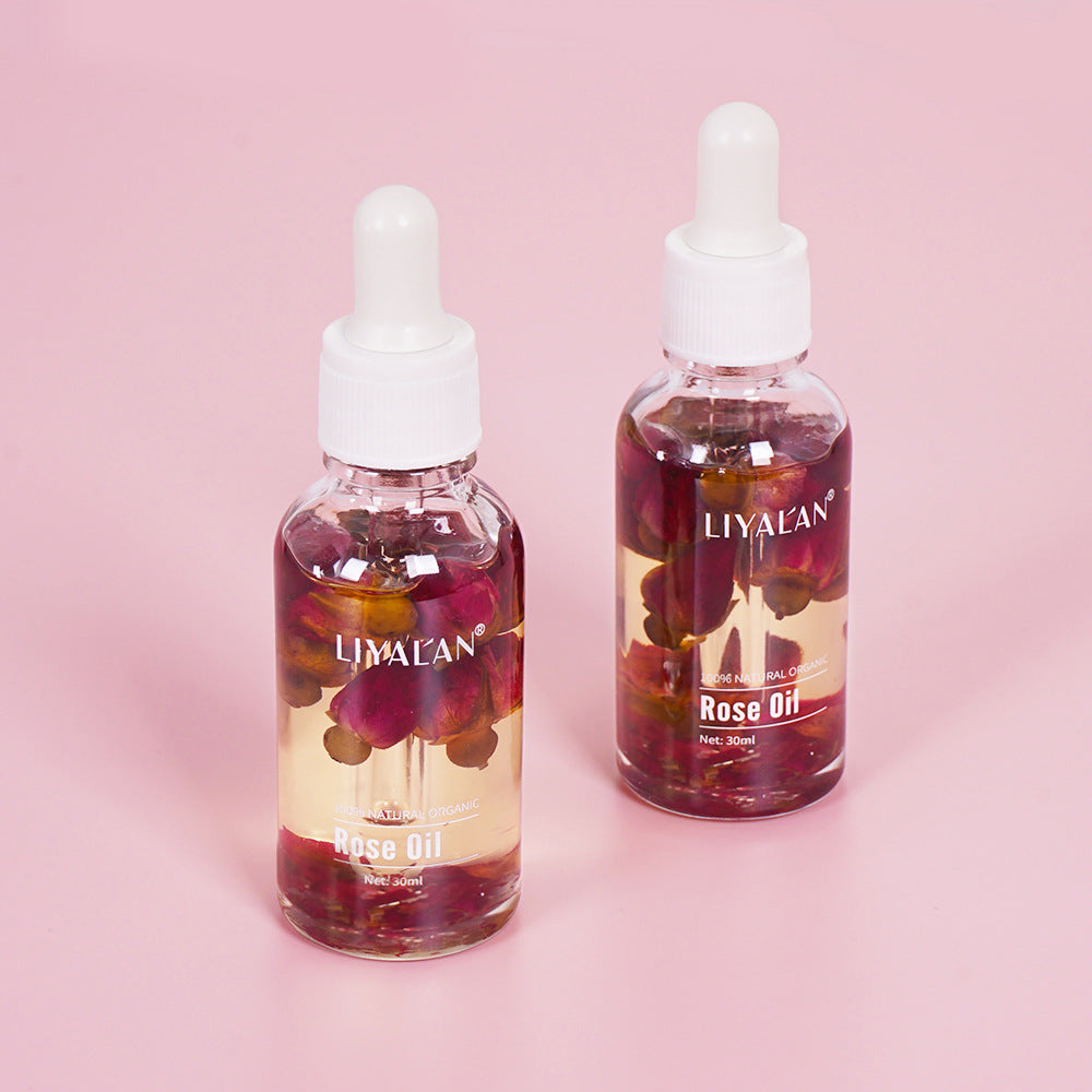 Product bottle with text overlay stating 'Rose Oil 100% NATURAL ORGANIC FOR SKIN AND HAIR' alongside fresh pink rose flowers, on soft lavender background