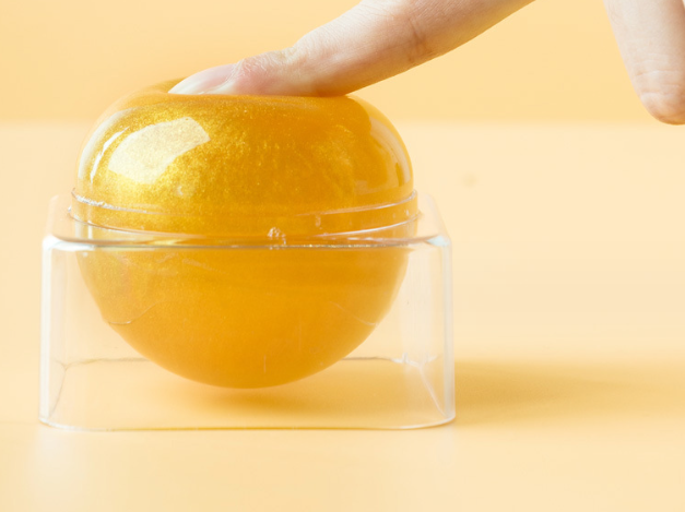 Close-up of hands holding transparent gel sphere with rotating band closure, showing crystal-clear golden formulation